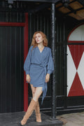 A woman with red hair stands outdoors in front of a black wooden building with red trim, wearing a blue and white gingham wrap mini dress with long sleeves and tan knee-high boots.