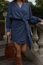 A woman wearing a blue and black gingham wrap dress with balloon sleeves, walking down outdoor stone steps while holding a brown fringe bucket bag.