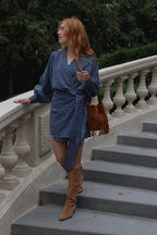 A woman with red hair stands on outdoor stone stairs wearing a blue and white gingham wrap dress, tan knee-high boots, and carrying a brown fringe shoulder bag.