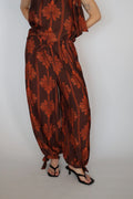 A person wearing a brown and rust-orange patterned harem jumpsuit with a drawstring waist, paired with black thong sandals, photographed against a plain light gray background.