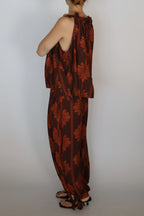 A woman shown from the back wearing a brown and rust-orange patterned halter jumpsuit with wide-leg pants and black strappy sandals, standing against a plain light gray background.