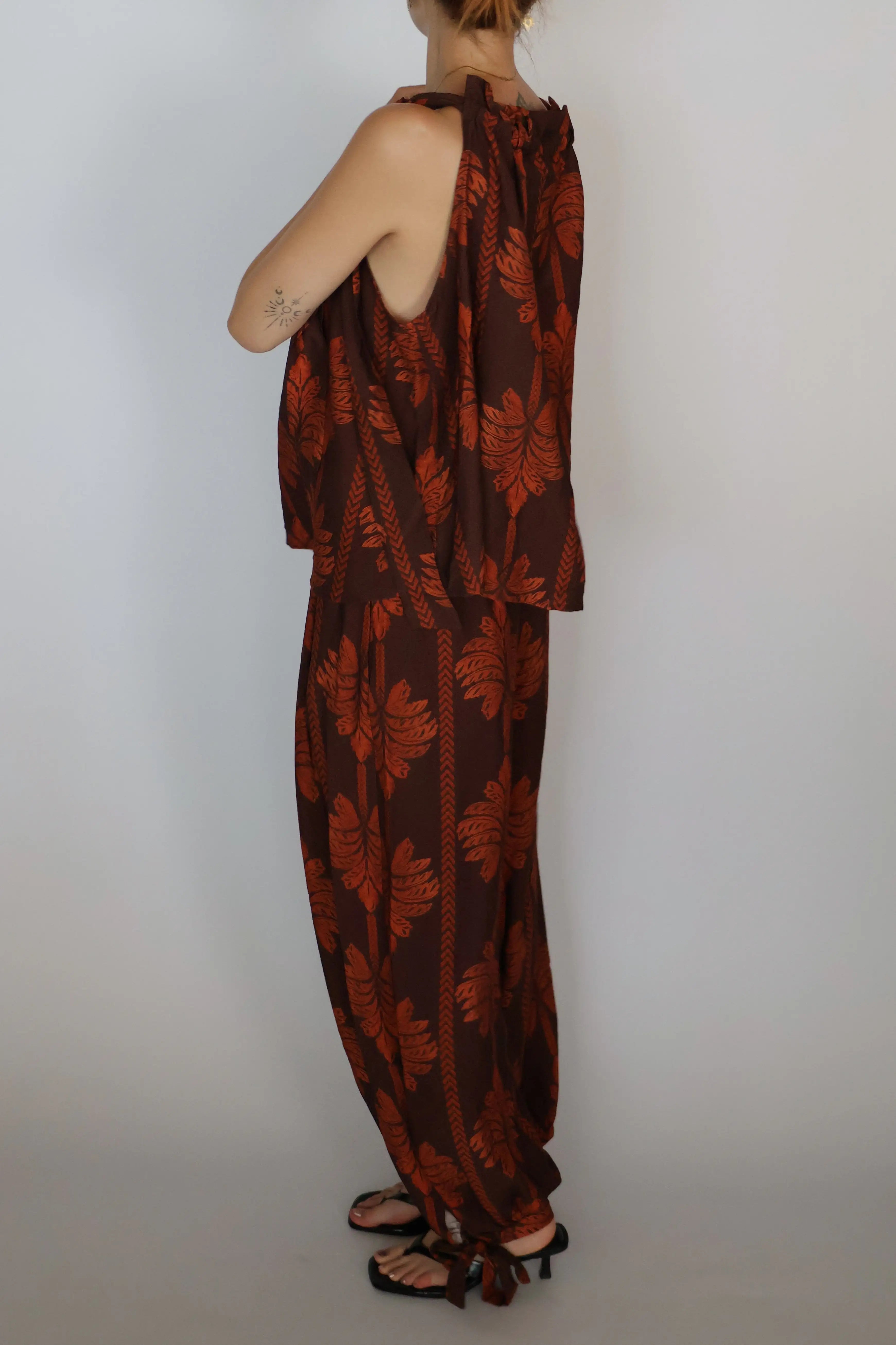 A woman shown from the back wearing a brown and rust-orange patterned halter jumpsuit with wide-leg pants and black strappy sandals, standing against a plain light gray background.