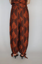 A person viewed from the back wearing loose brown harem-style pants with a rust-orange palm leaf and vertical stripe pattern, paired with black heeled sandals against a plain light gray background.