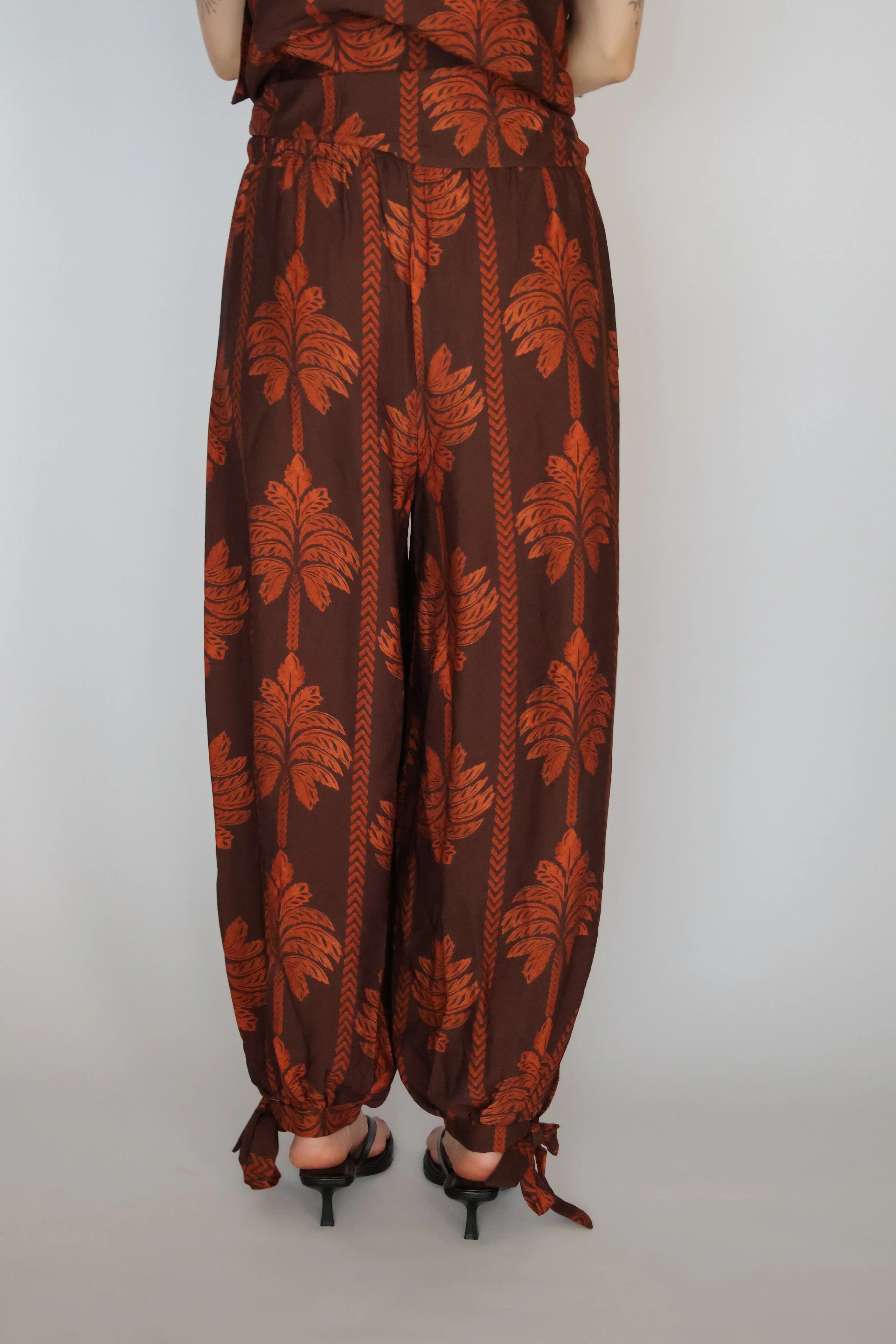 A person viewed from the back wearing loose brown harem-style pants with a rust-orange palm leaf and vertical stripe pattern, paired with black heeled sandals against a plain light gray background.
