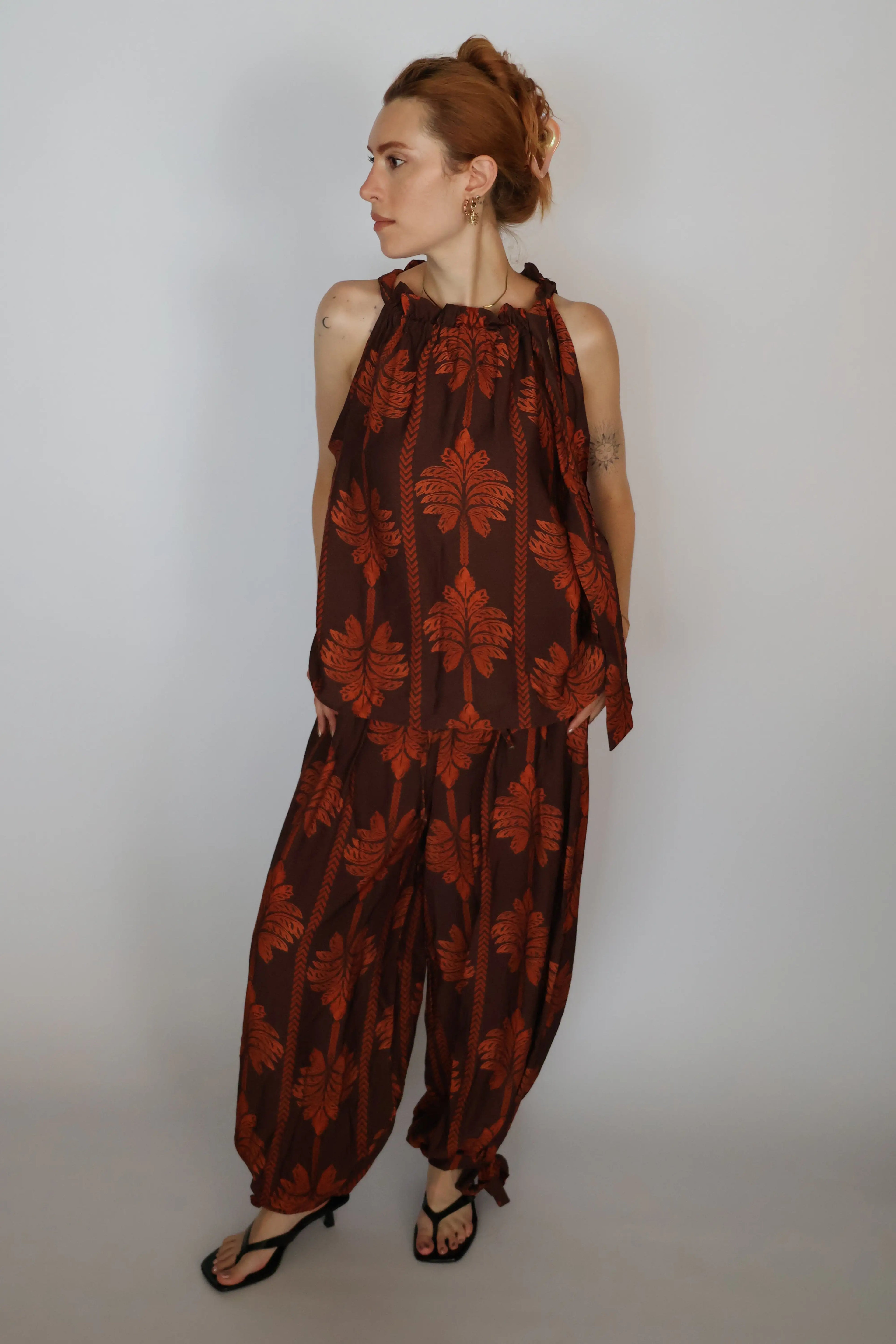 A woman modeling a sleeveless brown and rust-orange patterned jumpsuit with a halter neckline and wide-leg harem-style pants, standing against a plain light gray studio background.