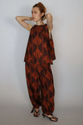 A woman modeling a brown and rust-orange patterned two-piece outfit consisting of a sleeveless top and matching wide-leg pants, paired with black strappy heels, against a plain light gray studio background.