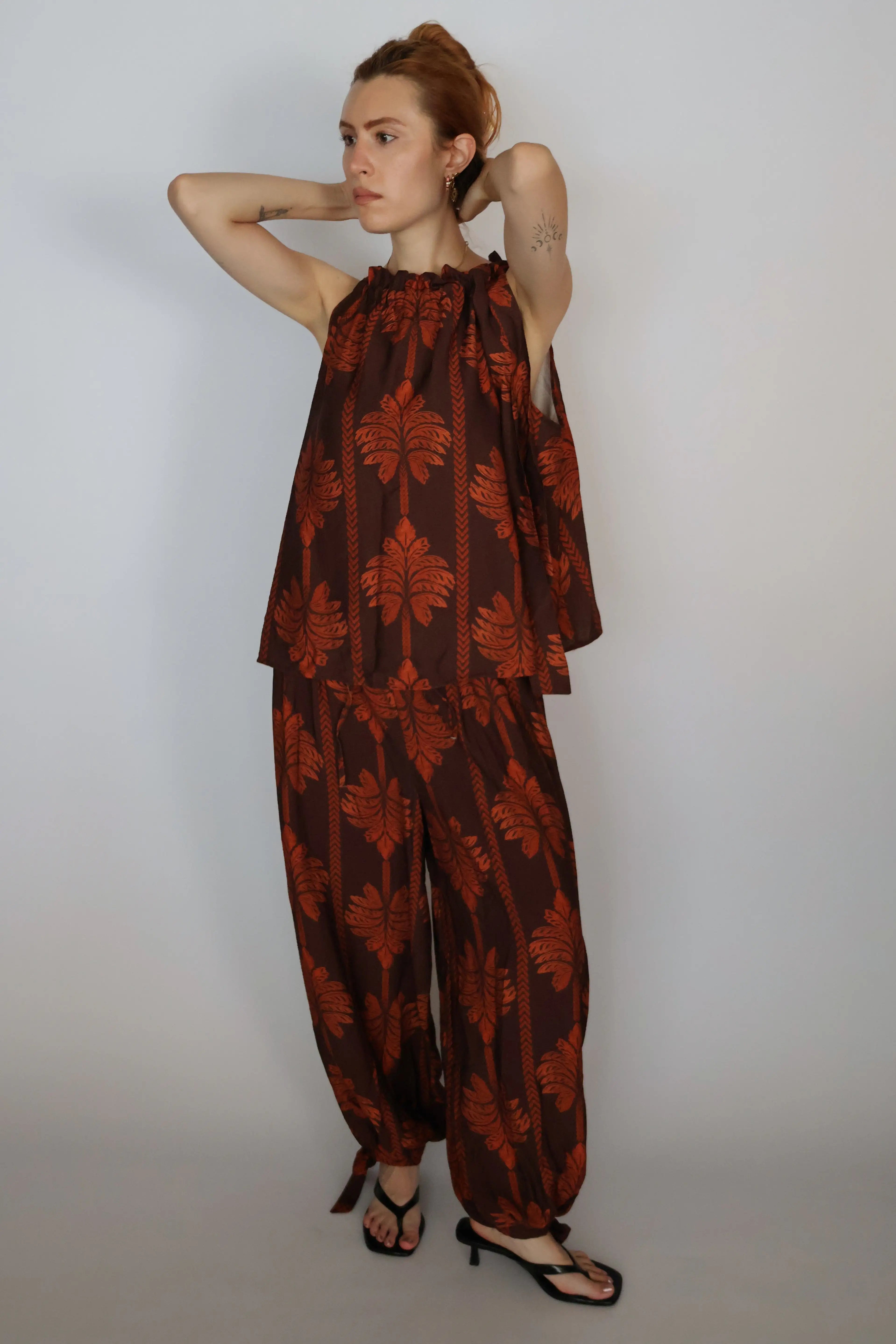 A woman modeling a brown and rust-orange patterned two-piece outfit consisting of a sleeveless top and matching wide-leg pants, paired with black strappy heels, against a plain light gray studio background.