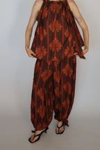 A model wearing a brown and rust-orange patterned two-piece set consisting of a sleeveless halter top and matching wide-leg pants, paired with black sandals, against a plain light gray background.