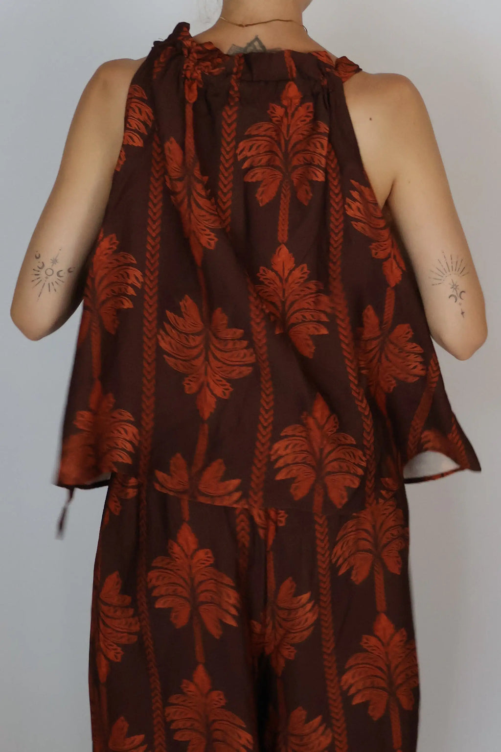 A sleeveless brown jumpsuit with a rust-orange palm leaf pattern, shown on a mannequin from the back against a plain light gray background.