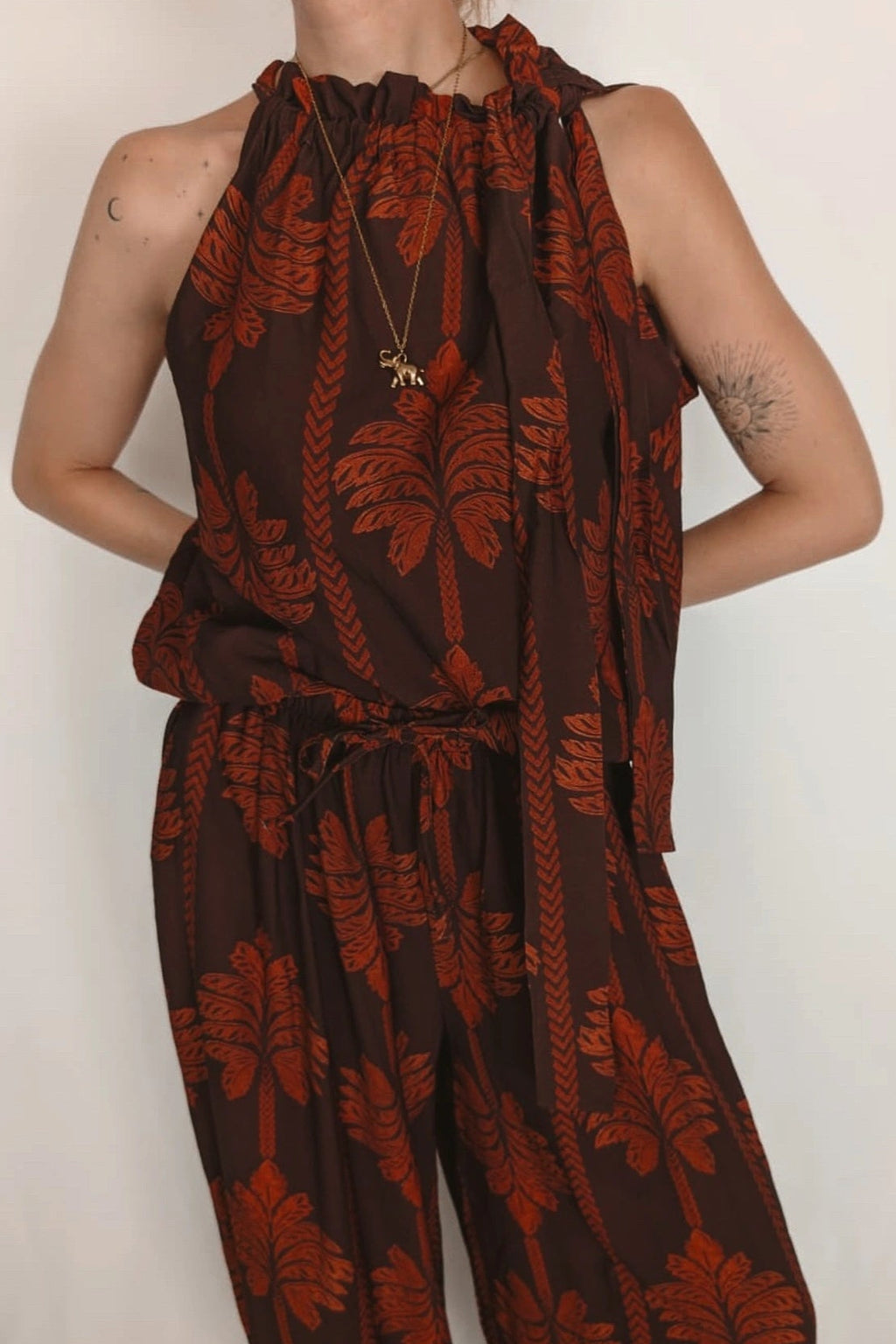 A sleeveless brown jumpsuit with a rust-orange palm leaf pattern, featuring a halter neckline, drawstring waist, and wide legs, shown on a model against a plain off-white background.