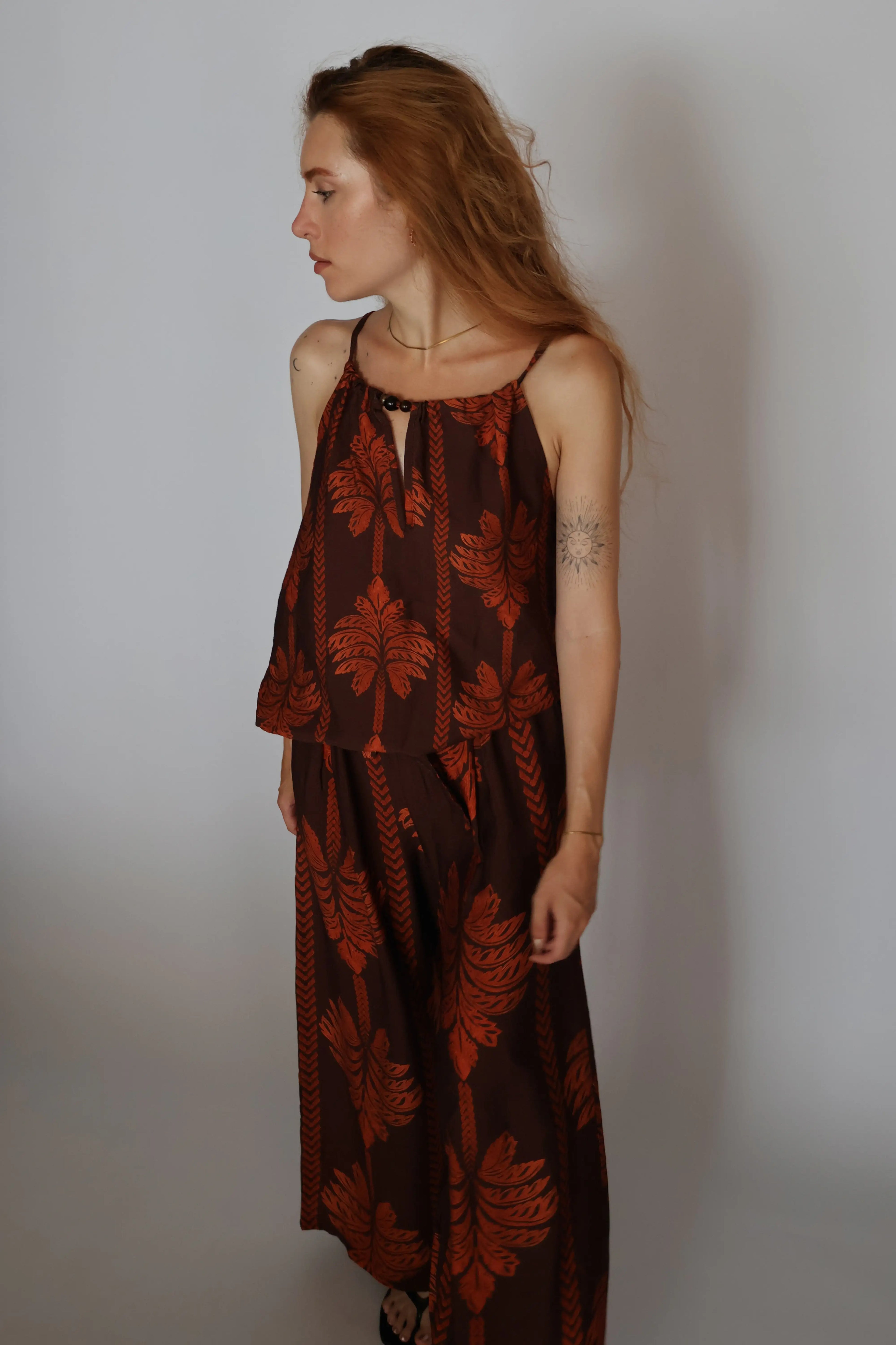 A woman with long light brown hair is shown in profile wearing a sleeveless brown dress with a rust-orange botanical leaf pattern, standing against a plain light gray wall.