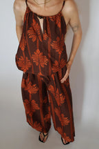 A woman modeling a brown and rust-orange tropical leaf print two-piece set consisting of a halter top and matching wide-leg pants, paired with black strappy heels against a plain light gray background.