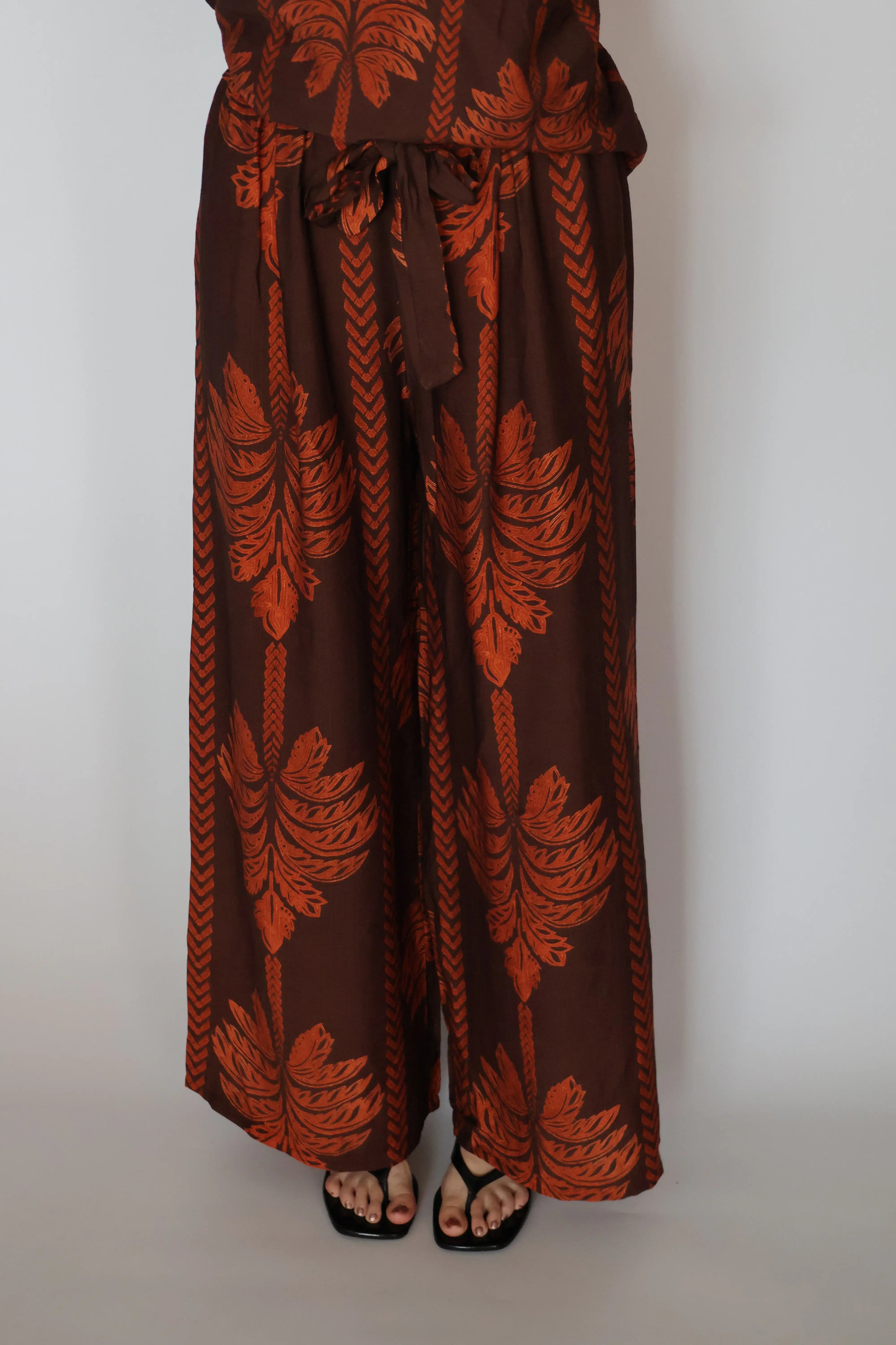A person wearing wide-leg brown pants with a rust-orange botanical leaf pattern, paired with black sandals, shown from the waist down against a plain light gray background.