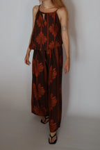 A woman modeling a sleeveless brown jumpsuit with a rust-orange botanical leaf pattern, paired with black sandals, against a plain light gray studio background.