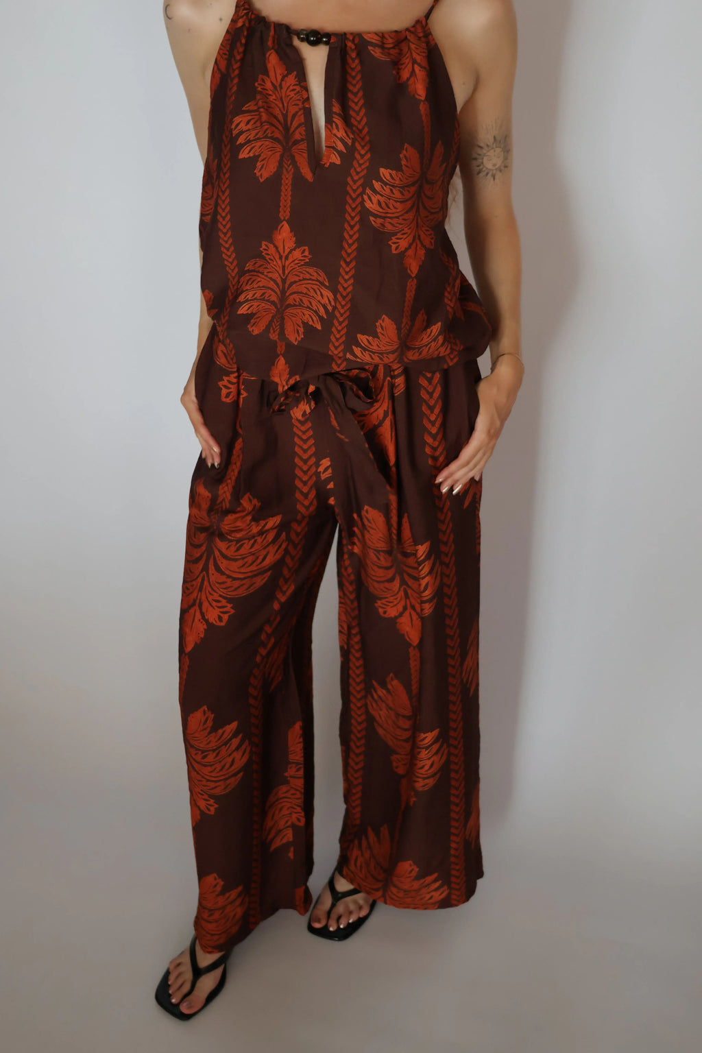 A woman wearing a brown halter jumpsuit with a rust-orange palm leaf pattern, paired with black flip-flop sandals, standing against a plain light gray background.