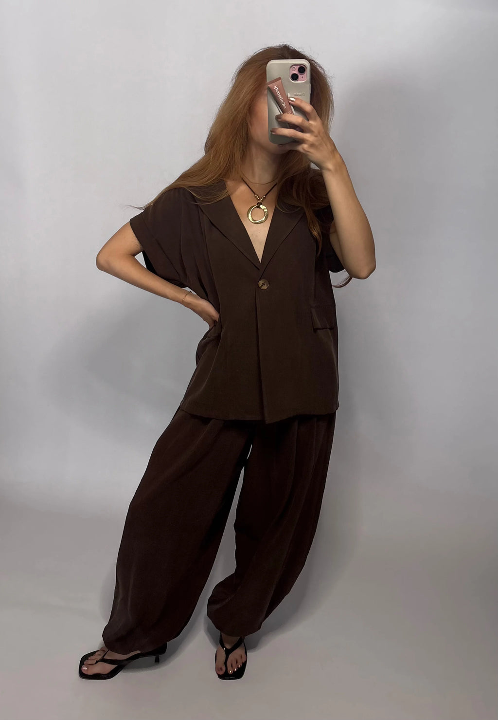 Woman with red hair in a brown two-piece set (ballon harem pants and short-sleeve gilet blazer) is walking towards a mirror with  phone in her hand