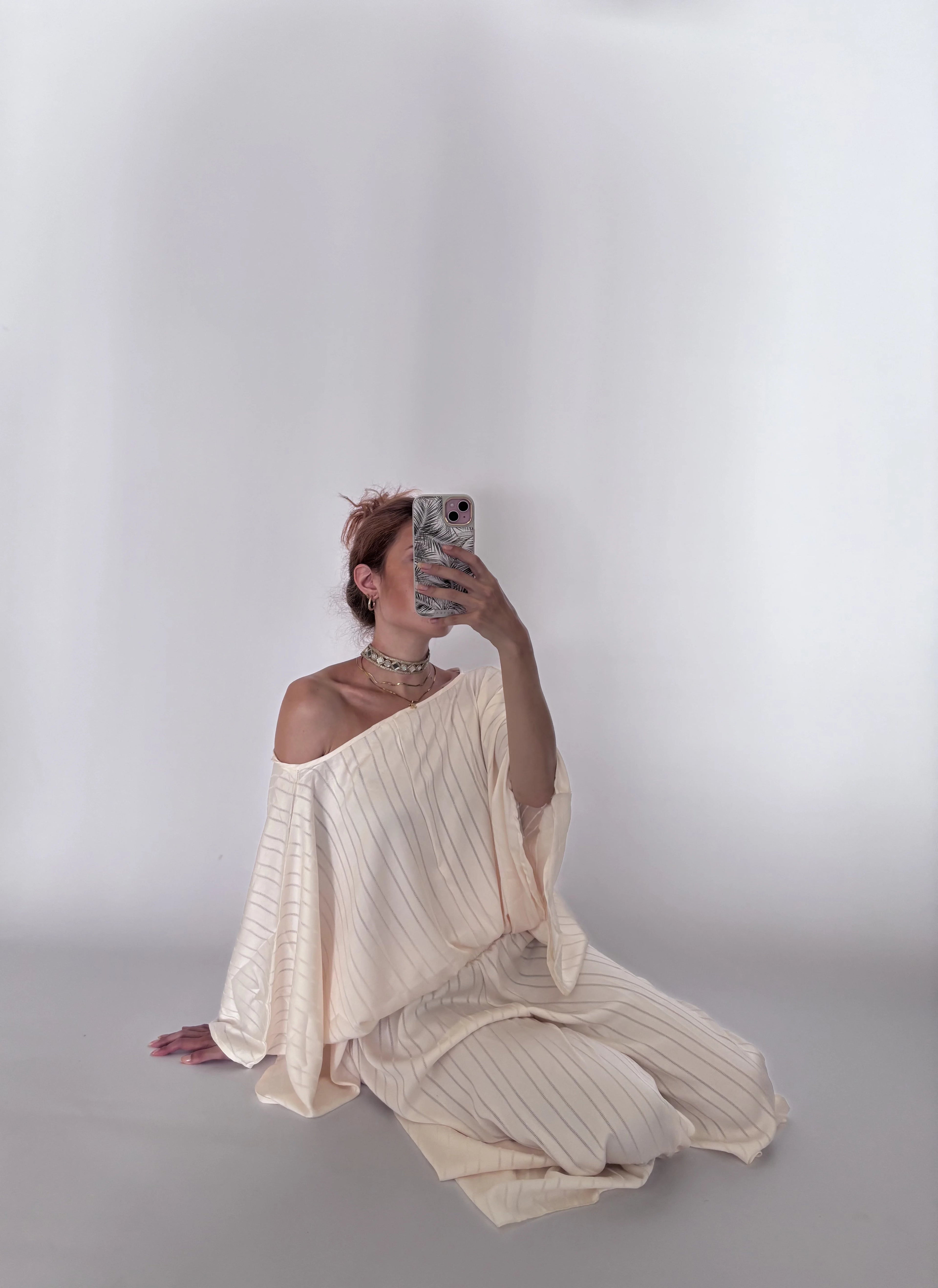 A woman sits on a light gray floor against a white studio backdrop, taking a mirror-style photo while wearing a loose, off-shoulder cream dress with thin vertical stripes.