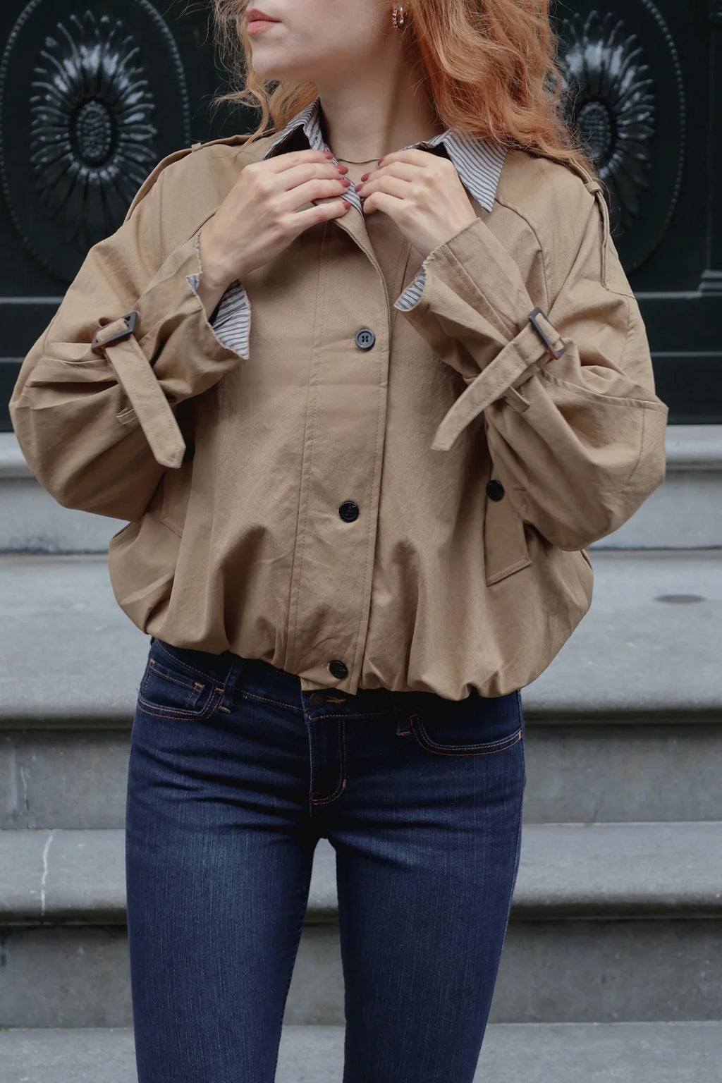 A woman wearing a cropped tan utility jacket with button-front closure and sleeve tab details, paired with dark blue jeans, standing outdoors in front of gray stone steps.
