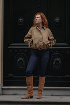 A woman with red hair stands in front of a large black ornate door wearing a cropped tan button-up shirt, dark blue jeans, and tan knee-high boots.