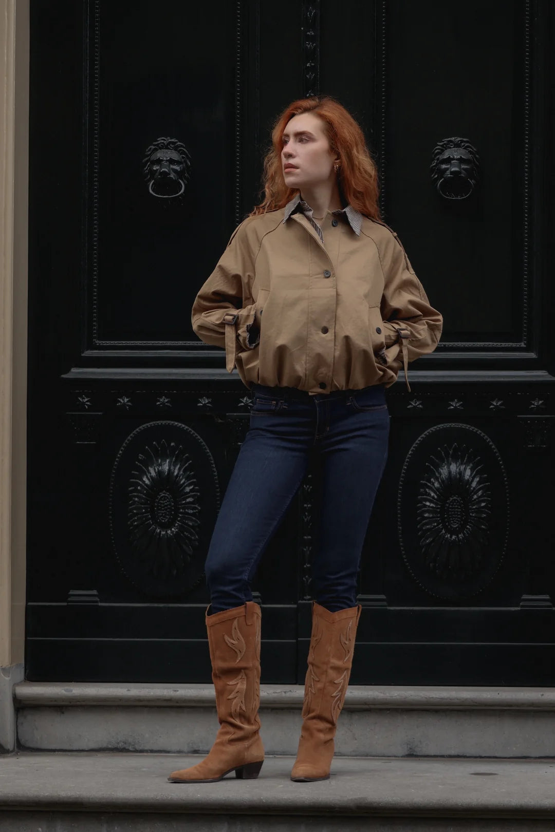 A woman with red hair stands in front of a large black ornate door wearing a cropped tan button-up shirt, dark blue jeans, and tan knee-high boots.