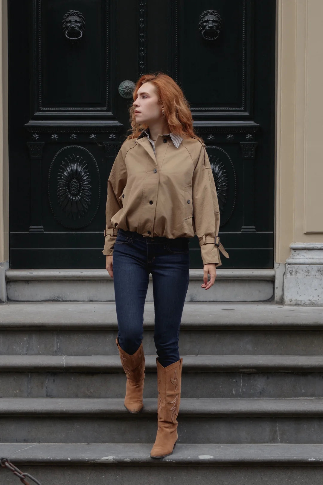 A woman with red hair walking down stone steps in front of a large black door, wearing a tan button-up shirt, dark blue jeans, and brown suede boots.
