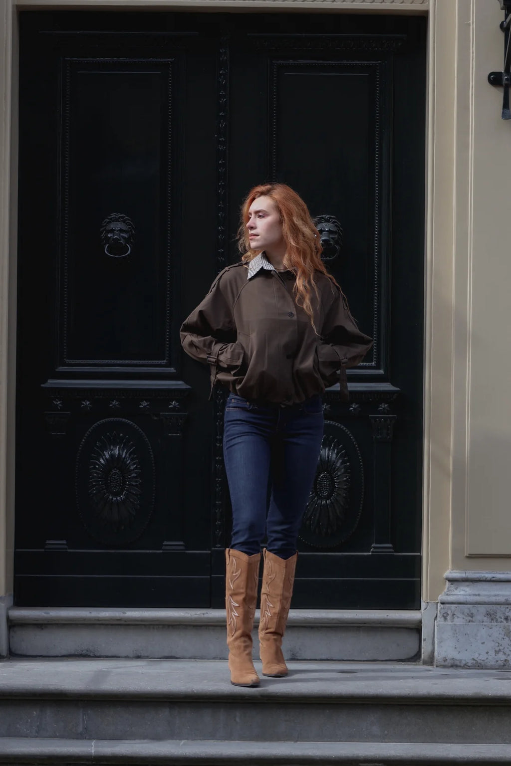 A woman with long red hair stands on stone steps in front of a large black door, wearing a dark brown long-sleeve top, dark blue jeans, and tan knee-high boots.