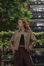 A woman with red hair stands outdoors wearing a tan utility-style jacket over a dark top and dark brown wide-leg pants, with trees and a bridge in the background.