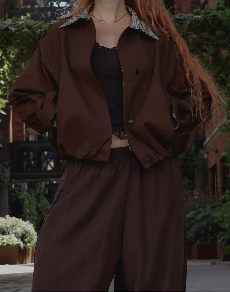 A person wearing a dark brown lightweight jacket with a striped collar, a black camisole, and matching dark brown wide-leg pants, standing outdoors in a garden setting.