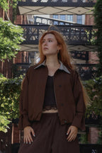 A woman with long red hair stands outdoors wearing a brown cropped jacket with a gray striped collar, a black top, and brown wide-leg pants, set against a background of brick buildings, greenery, and a black metal bridge.