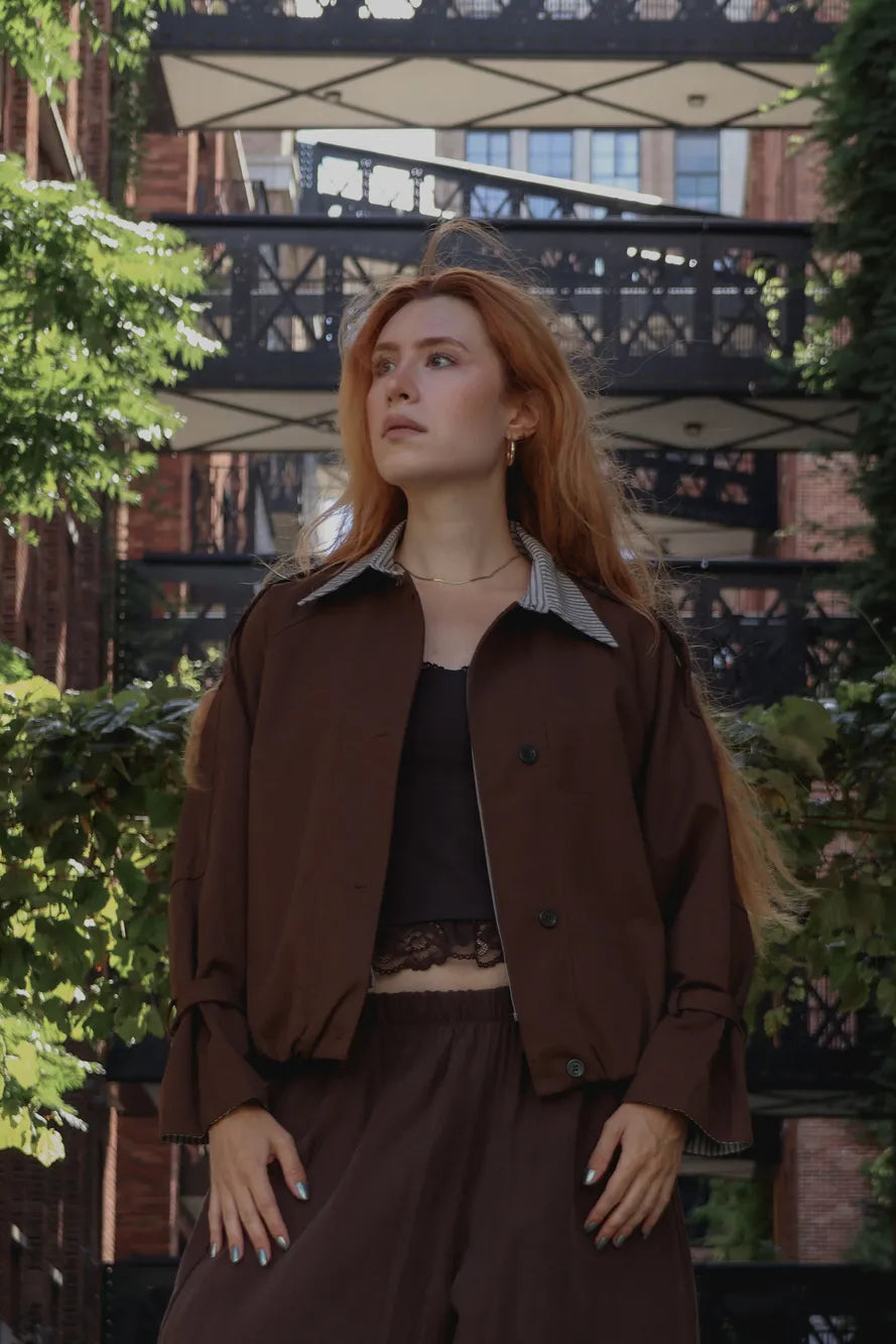 A woman with long red hair stands outdoors wearing a brown cropped jacket with a gray striped collar, a black top, and brown wide-leg pants, set against a background of brick buildings, greenery, and a black metal bridge.