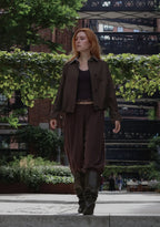 A woman walking outdoors in a dark brown shirt-jacket, matching loose pants, a black crop top, and dark boots, with greenery and a brick building in the background.