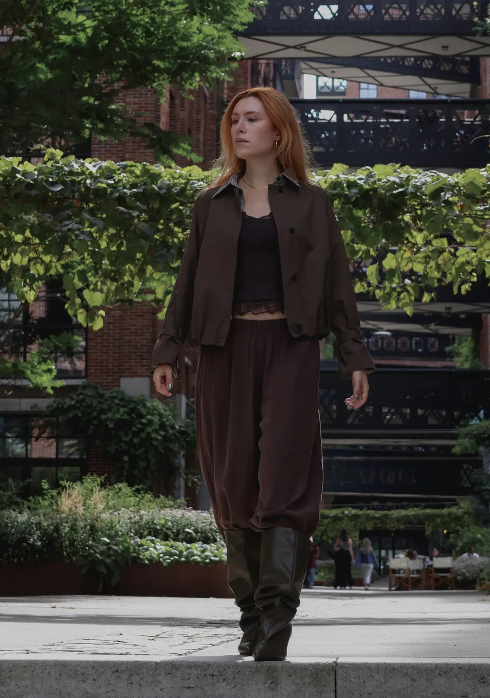 A woman walking outdoors in a dark brown shirt-jacket, matching loose pants, a black crop top, and dark boots, with greenery and a brick building in the background.