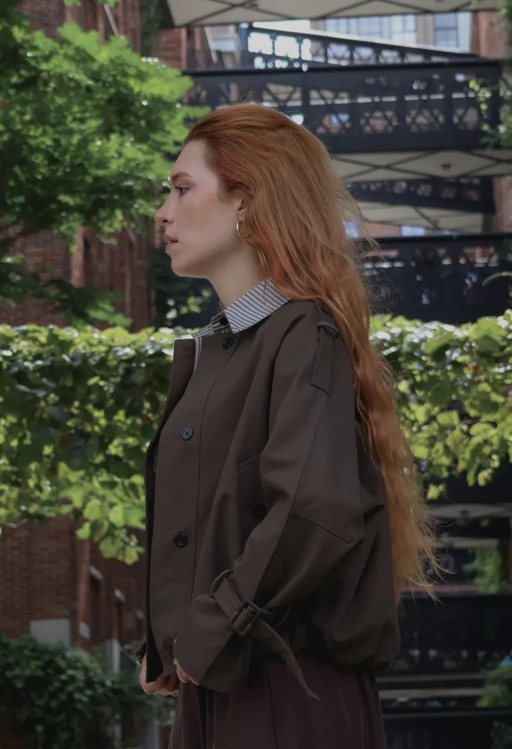 A woman with long red hair stands outdoors in profile, wearing a dark brown trench coat over a striped shirt and dark pants. The background features green foliage and a brick industrial building.