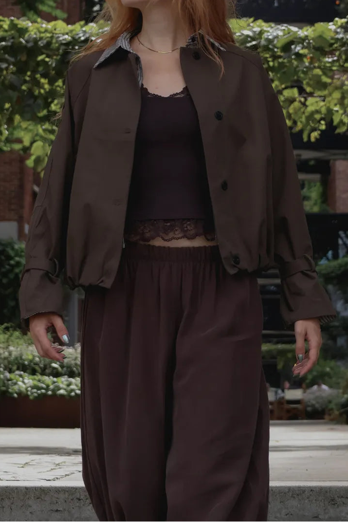 A woman with red hair stands outdoors wearing a dark brown cropped jacket, matching dark brown pants, and a dark brown camisole top. The background features brick buildings, greenery, and a black metal bridge.