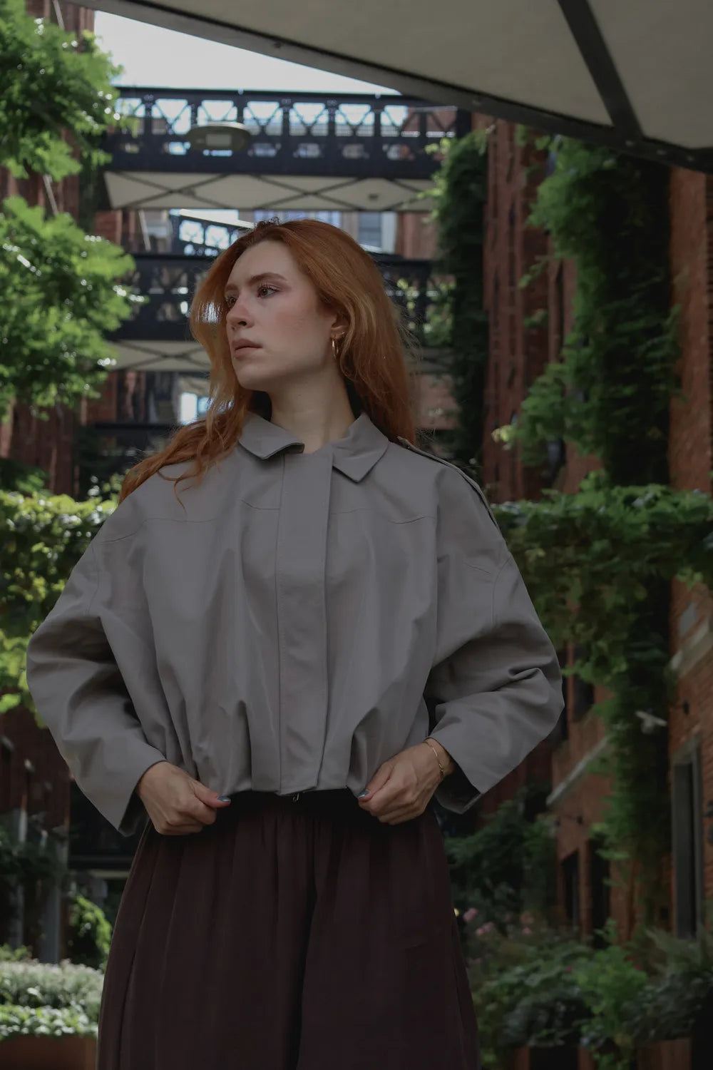 A woman with red hair wearing a light gray oversized long-sleeve jacket and a dark brown skirt, standing in an outdoor urban alleyway with brick buildings and greenery.