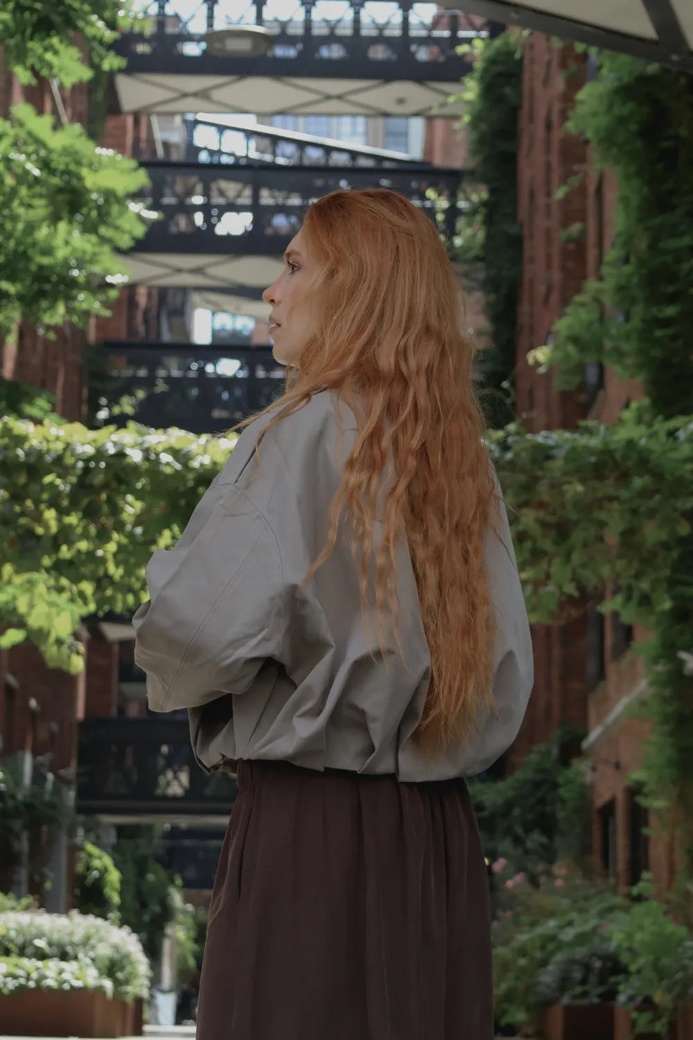 A woman with long, wavy strawberry blonde hair is shown from the back, wearing a loose, oversized light gray top and a dark brown pants, standing in an outdoor urban alleyway with brick buildings and greenery.