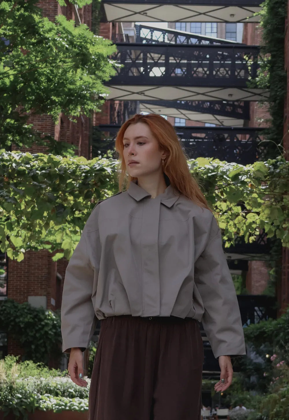 A woman with red hair wearing a light gray cropped utility jacket and dark brown wide-leg pants, standing in an outdoor garden setting with green foliage and a brick building in the background.
