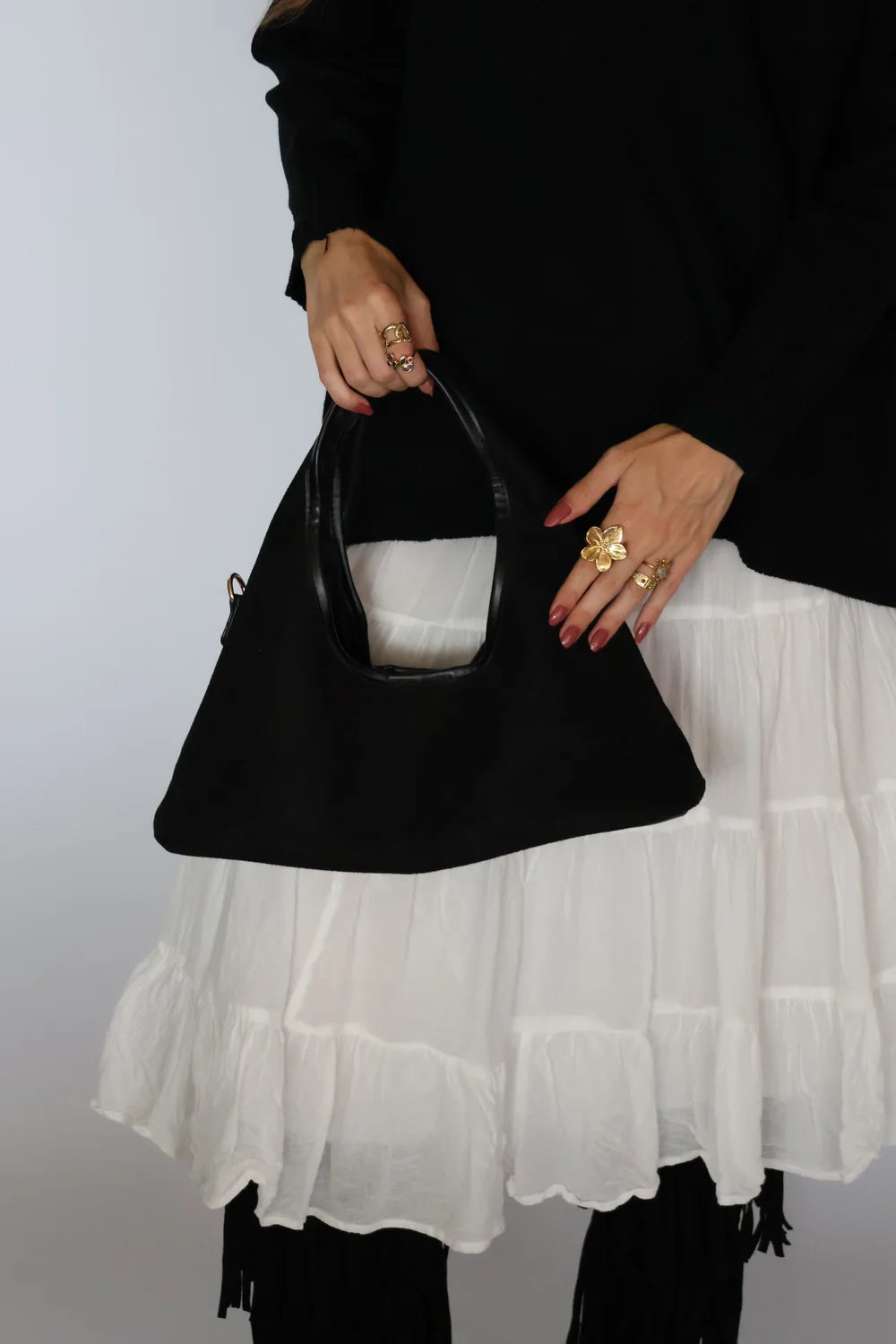 A person wearing a black long-sleeve top and a white tiered skirt holds a black structured handbag with a top handle against a plain light gray background.