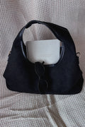 A black suede shoulder bag with black leather trim, shown on a textured beige waffle-knit blanket. A white book and black sunglasses are placed inside the bag as styling props.